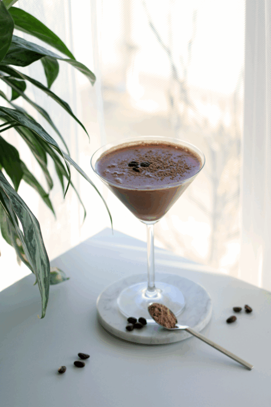 Cremiger Mühltini - alkoholfreie Espresso Martini-Alternative in einem Martini-Glas, garniert mit Kaffeebohnen und CHOCO CHARME Bio Vegan Protein Pulver auf einem weißen Marmor-Untersetzer neben einer Zimmerpflanze.