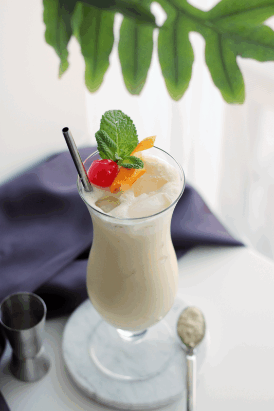 Exotischer Schalcolada Cocktail mit GOLDEN GLOW Bio Vegan Protein Mix in einem Hurricaneglas mit Schaumkrone, garniert mit Minze, Kirsche und Orangenschale auf einem Marmor-Untersetzer.