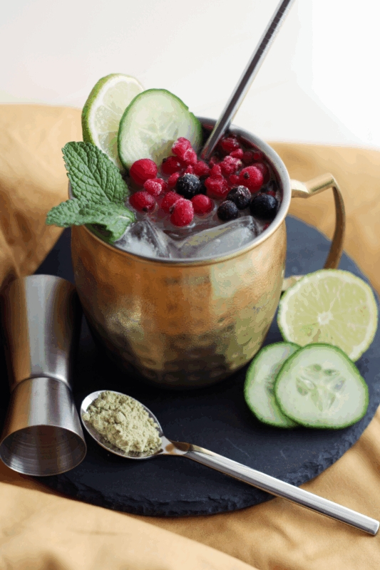 Erfrischender Schalk Mule in einem Kupferbecher, garniert mit gefrorenen Beeren, Gurkenscheiben und Minze, serviert auf einer Schieferplatte mit einem Löffel AMINO POWER Bio Vegan Protein Mix.