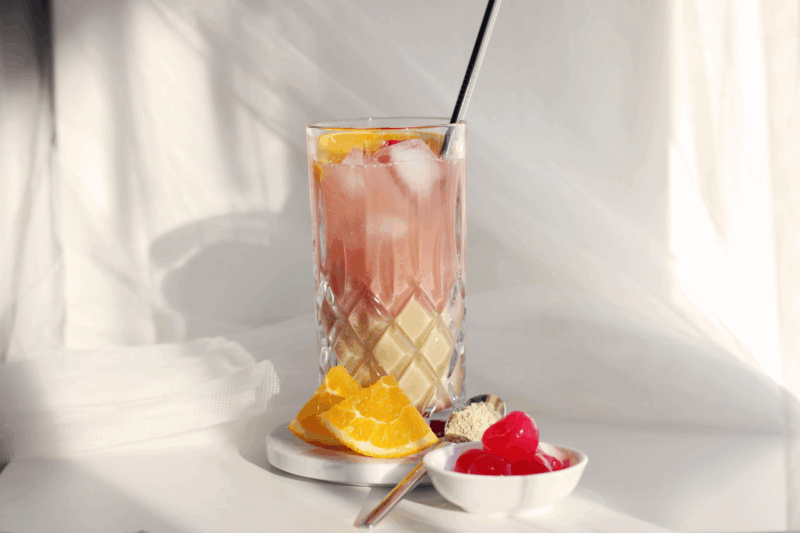 Erfrischender Schalk on the Beach Cocktail mit TROPICAL TREASURE Bio Vegan Protein in einem Kristallglas mit Eiswürfeln, Orangenscheiben und Kirschen auf hellem Hintergrund.