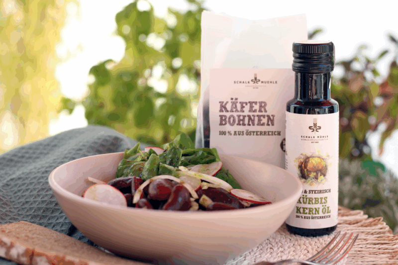 Steirischer Käferbohnensalat mit Vogerlsalat in einer Schüssel mit Schalk Mühle Bio Käferbohnen und Schalk Mühle Bio steirischem Kürbiskernöl im Hintergrund