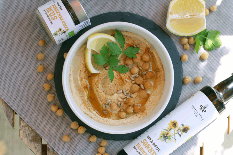 Sonnenblumen-Hummus auf grauem Tischtuch, mit Kichererbsen, Sonnenblumenkerne, Zitronenspalte und Petersilie garniert - mit Schalk Mühle Sonnenblumenkernmus und Sonnenblumenöl