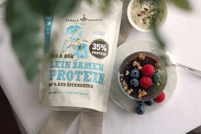 Leinsamen Protein Pudding angerichtet auf weißem Untergrund, mit Marmorplatte, frischen Beeren, Walnüssen und Schlagobers, daneben Schalk Mühle Leinsamen Protein Pulver.