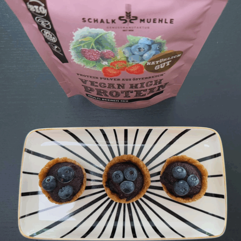 Vegane Beeren-Protein-Törtchen mit proteinreicher Beerenfüllung auf Dattel-Cashew-Boden, dekoriert mit frischen Blaubeeren – angerichtet auf schwarz-weißem Teller mit Schalk Mühle Multi Beeren Protein Mix Beutel im Hintergrund.