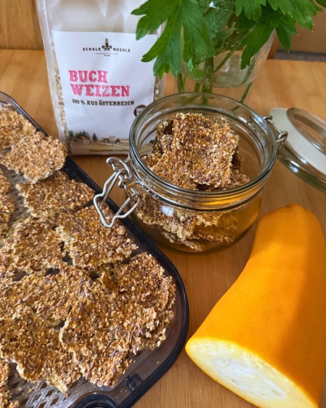 Selbstgemachte roh-vegane Zucchini-Cracker mit gekeimtem Buchweizen und Leinsamen auf einem Dörrgitter und in einem Glasbehälter, daneben gelbe Zucchini, ein Bund frische Petersilie und eine Packung Bio-Buchweizen von Schalk Mühle.