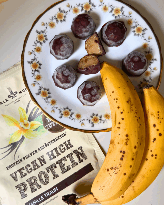 Vegane Monkey-Chocolates auf einem Blumenteller, daneben reife Bananen und Bio Vanille Protein Pulver Mix der Schalk Mühle.