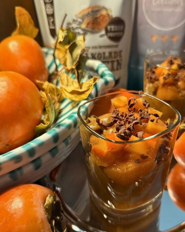Veganer Schalkoladepudding mit Kakicreme im Glas, getoppt mit frischen Kakistücken und Schokoraspeln, daneben reife Kakis und Schalkolade Proteinpulver der Schalk Mühle.