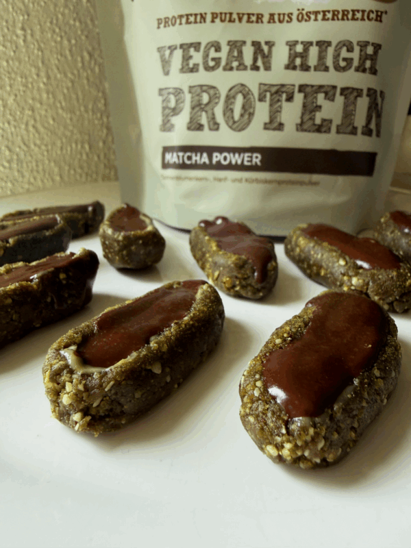 Mehrere Hanf-Matcha-Stixmit dunkler Schokoglasur auf der Oberseite sind auf einer hellen Fläche aufgereiht. Im Hintergrund steht eine Packung „Vegan High Protein Matcha Power“ Pulver der Marke Schalk Mühle.
