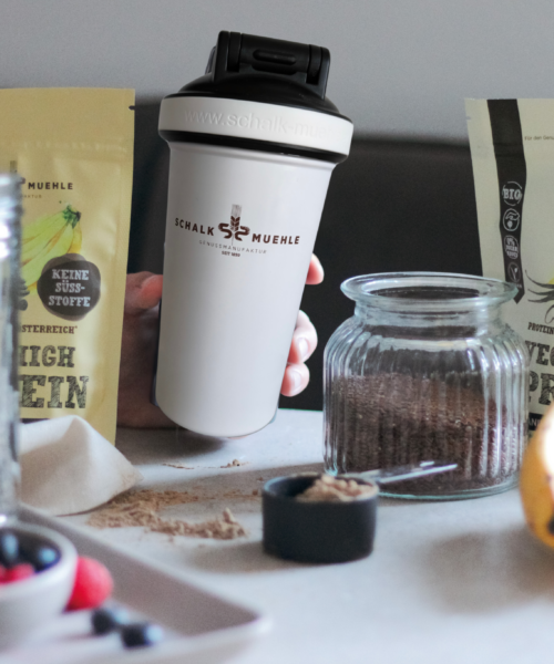 Am Bild zwei Protein Pulver (Kakao Banane und Vanille) der Schalk Mühle, in der Mitte der Schalk Mühle Shaker, davor Bananen, Leinsamen in einem Glas, links vorne Beeren auf einem kleinen Schälchen, und dahinter ebenfalls Samen