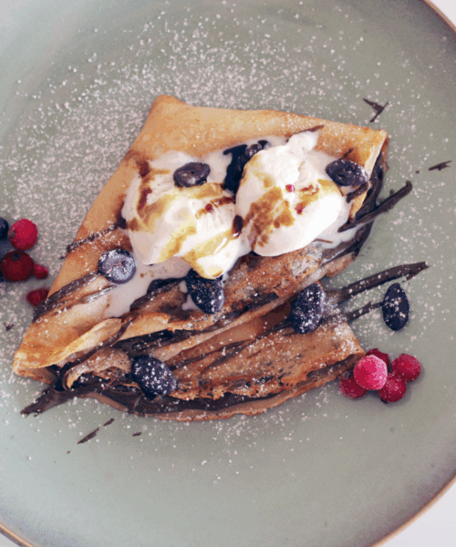 Eine goldbraun gebackene, gefaltete Palatschinke auf einem hellblauen Teller, gefüllt mit Schokoladencreme und getoppt mit zwei Kugeln Vanilleeis. Das Dessert ist mit Schokoladensauce garniert, mit Puderzucker bestäubt und mit gefrorenen Blaubeeren sowie roten Johannisbeeren dekoriert.