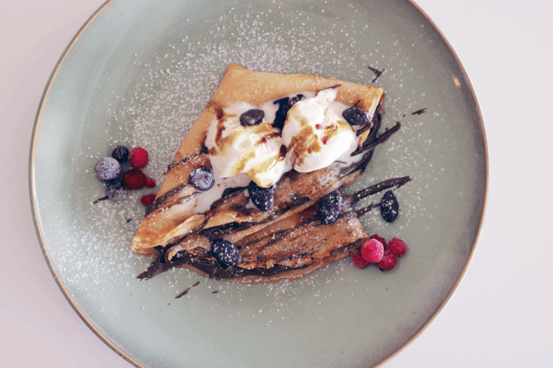 Eine goldbraun gebackene, gefaltete Palatschinke auf einem hellblauen Teller, gefüllt mit Schokoladencreme und getoppt mit zwei Kugeln Vanilleeis. Das Dessert ist mit Schokoladensauce garniert, mit Puderzucker bestäubt und mit gefrorenen Blaubeeren sowie roten Johannisbeeren dekoriert.