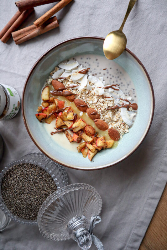 schön gestaltete Bowl, dekoriert mit Äpfel, Mandeln, Kokosflocken, Chia und Haferflocken, goldener Löffel und rohe Chiasamen daneben