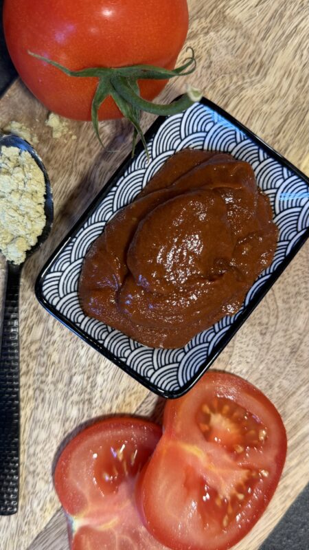 aus der Vogelperspektive die BBQ Sauce in einem kleinen Schälchen mit Muster, daneben eine ganze Tomate und Tomatenscheiben sowie ein Löffel mit dem Schalk Mühle Protein Pulver