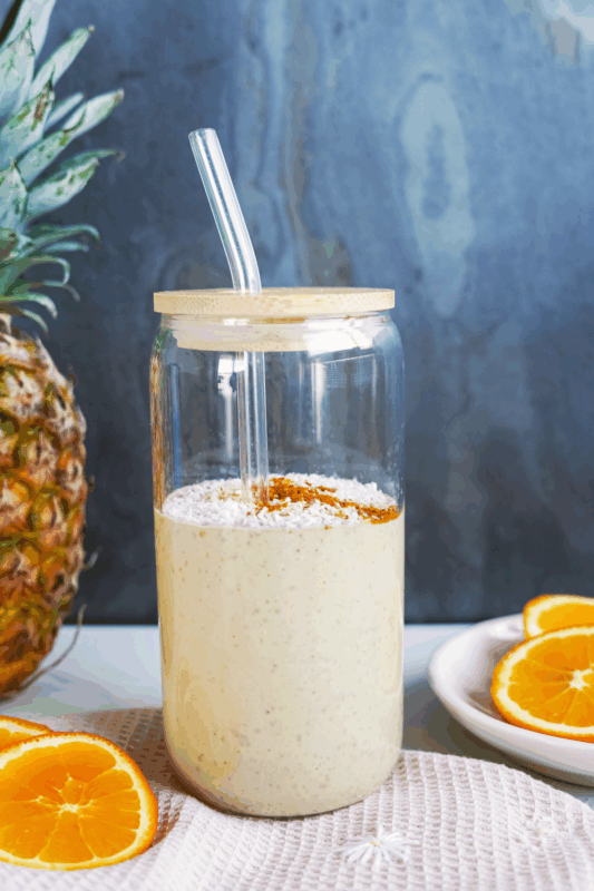 Cremiger Tropical Protein Milkshake mit Schalk Mühle Tropical Treasure Bio Proteinpulver in einem Glas mit Bambusdeckel, garniert mit Kokosraspeln und Zimt, neben frischen Orangenscheiben und einer Ananas.