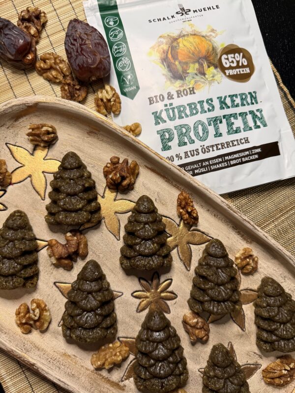 Vegane No-Bake Gingerbread Trees aus Datteln, Walnüssen und Kürbiskernprotein, weihnachtlich geformt und auf dekorativem Teller angerichtet, daneben Bio Kürbiskern Protein von der Schalk Mühle – gesunder Weihnachts-Snack ohne Backen.