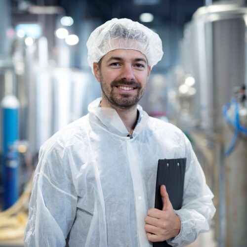 lächelnder Mann in einer Produktionshalle, mit Schutzmantel in weiß und Häubchen in weiß